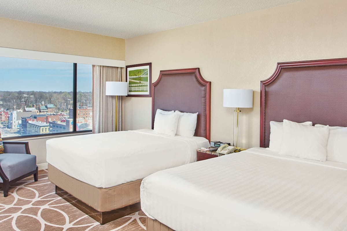 Quarto de hotel com duas camas, vista para a cidade, uma escrivaninha com luminária e uma cadeira.