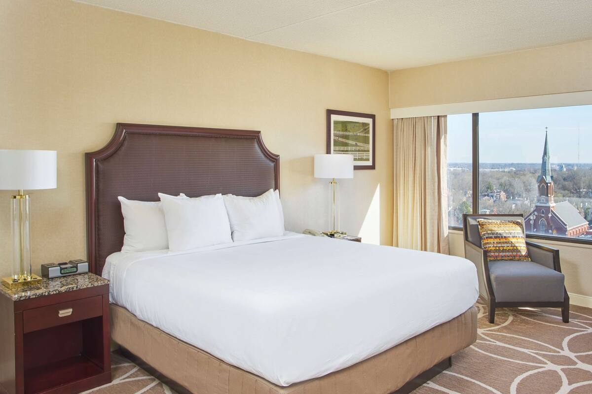 Quarto de hotel com uma cama grande, duas mesas de cabeceira com luminárias, uma poltrona e vista para a cidade através de uma janela ampla.
