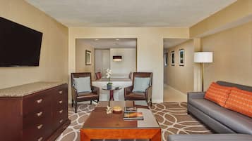 A hotel room with a sofa, a coffee table, a TV, a dresser, and a dining area with chairs and a table.