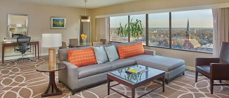 A hotel room with a large window offering a city view, a sofa with patterned cushions, a small table, and a desk with a chair.