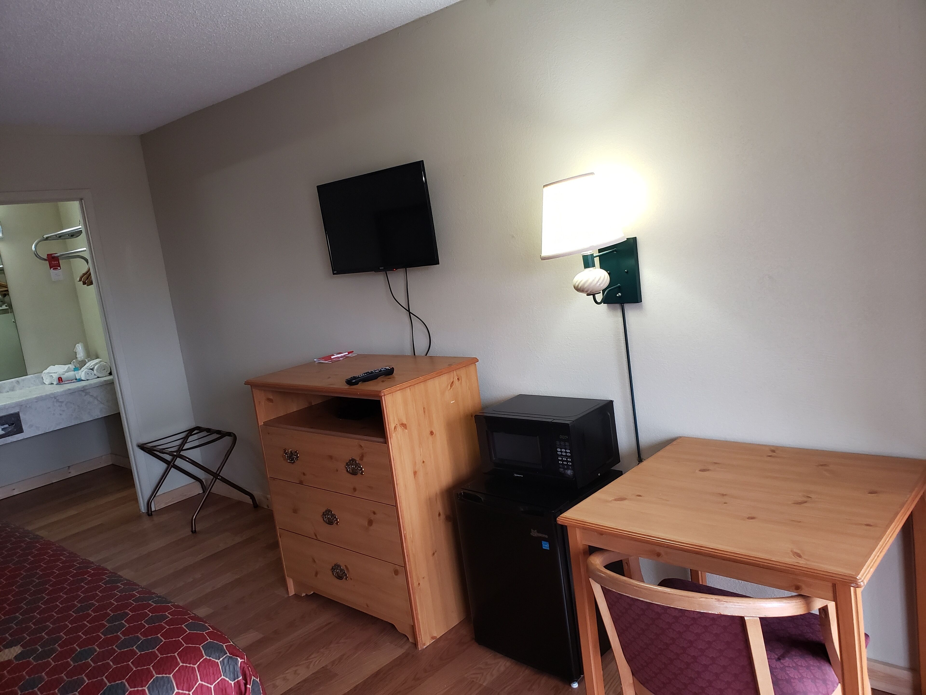 A compact room with a wooden desk, a small black refrigerator, a television mounted on the wall, and a bedside table with a mirror.