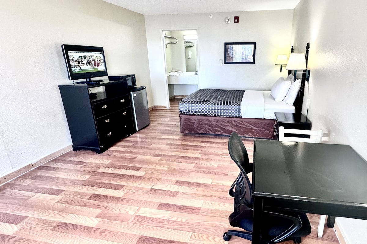 A compact hotel room with a bed, a desk, a chair, a TV, a dresser, and a bathroom visible in the background.