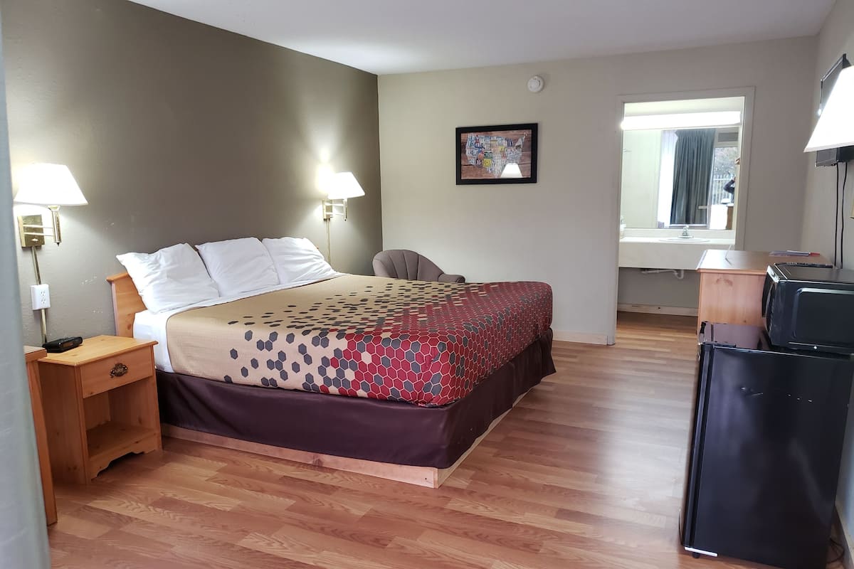 A hotel room with a bed, bedside lamps, a desk, a chair, a microwave, and a refrigerator.