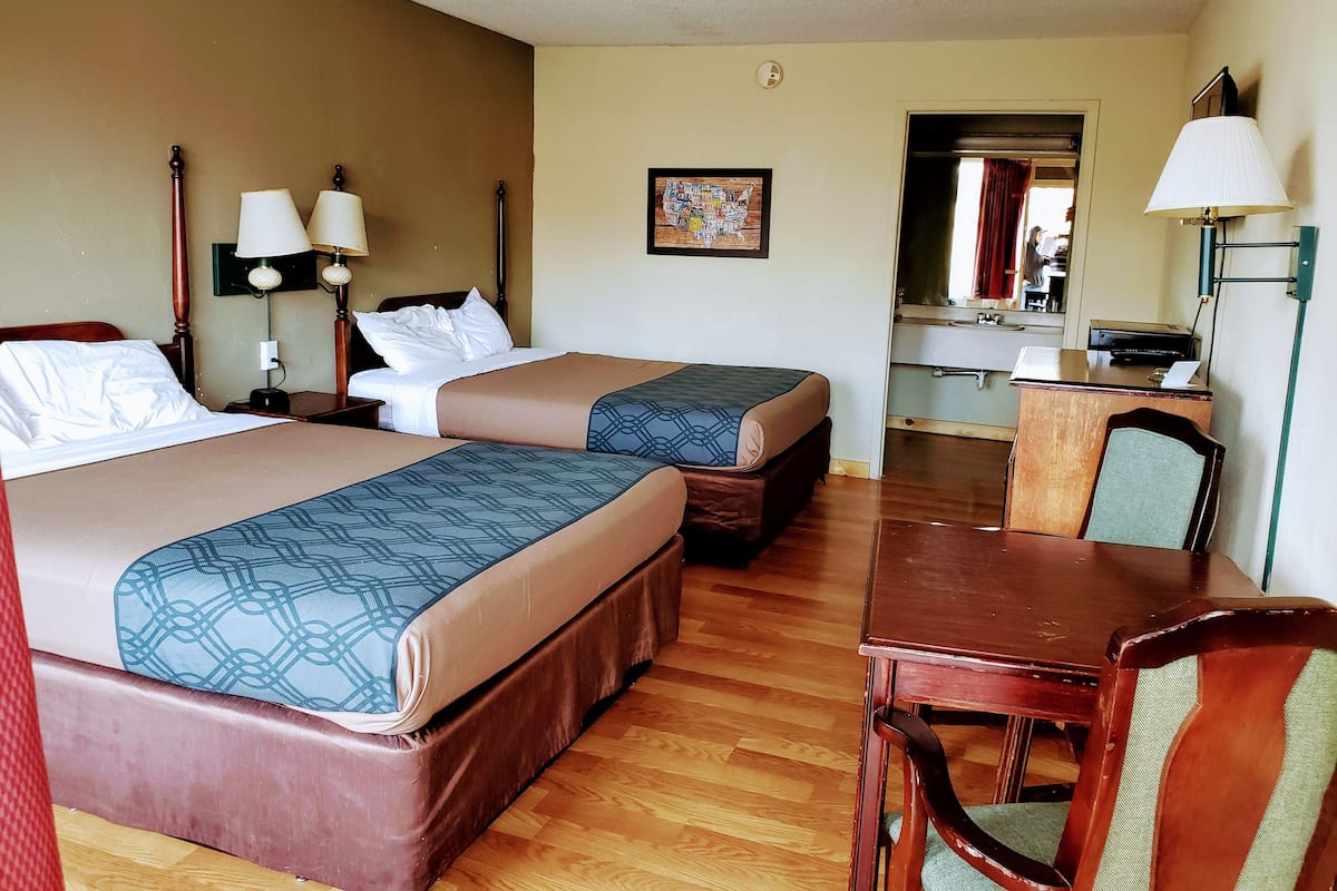 A hotel room with two beds, a desk, a chair, a desk lamp, a wall clock, and a bathroom visible through an open door.