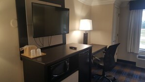 A hotel room with a flat-screen TV, a desk with a lamp, a microwave, a chair, and a window with curtains. - Regency inn and Suites (Pensacola)