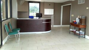 A reception area with a curved desk, a computer, a window with a view, a chair, and a small shelf with items. - Regency inn and Suites (Pensacola)