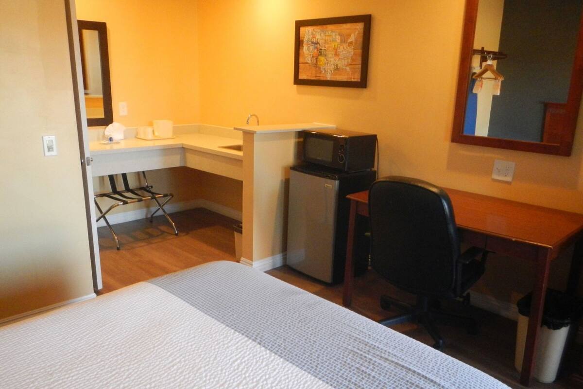 A compact hotel room with a bed, a small kitchenette, a desk, and a chair.