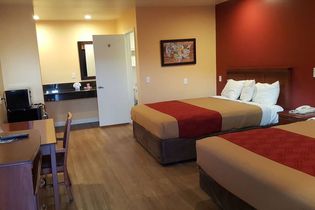 A hotel room with two beds, a desk, a chair, a small kitchenette, and a mirror.