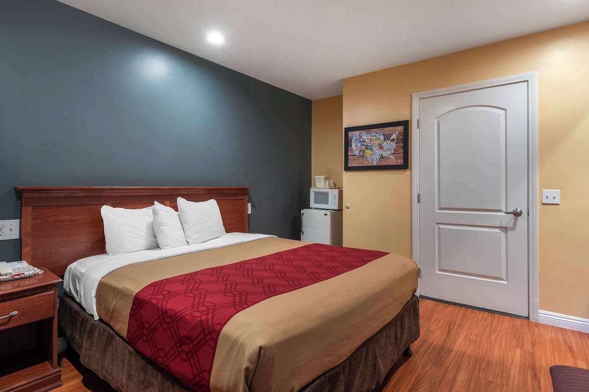A hotel room with a large bed, a wooden headboard, a red and beige bedspread, a white door, and a small white refrigerator.