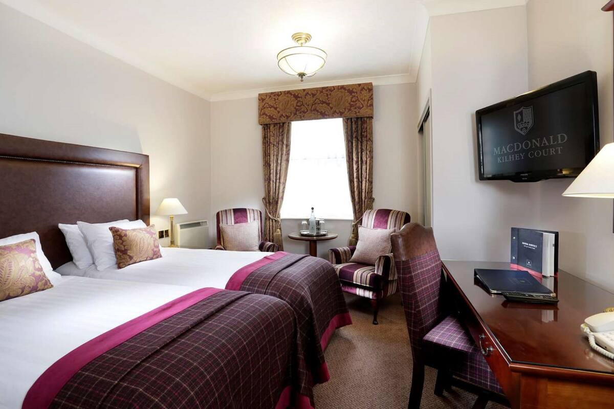 A hotel room with two beds, a desk, a chair, a television, and a window with curtains.