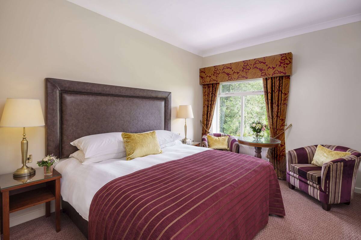 A hotel room with a large bed, two bedside tables with lamps, a chair, and a window with curtains.
