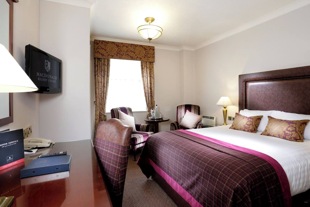 A hotel room with a bed, a desk with a telephone and a remote control, a chair, a small table with a lamp, and a television on the wall.