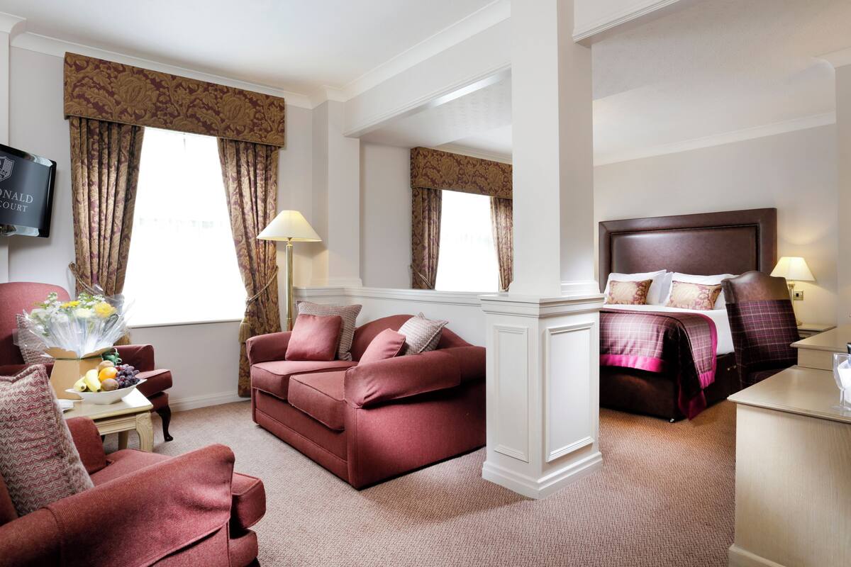 A hotel room with a red sofa, a bed with a headboard, a desk with a lamp, and a TV on the wall.