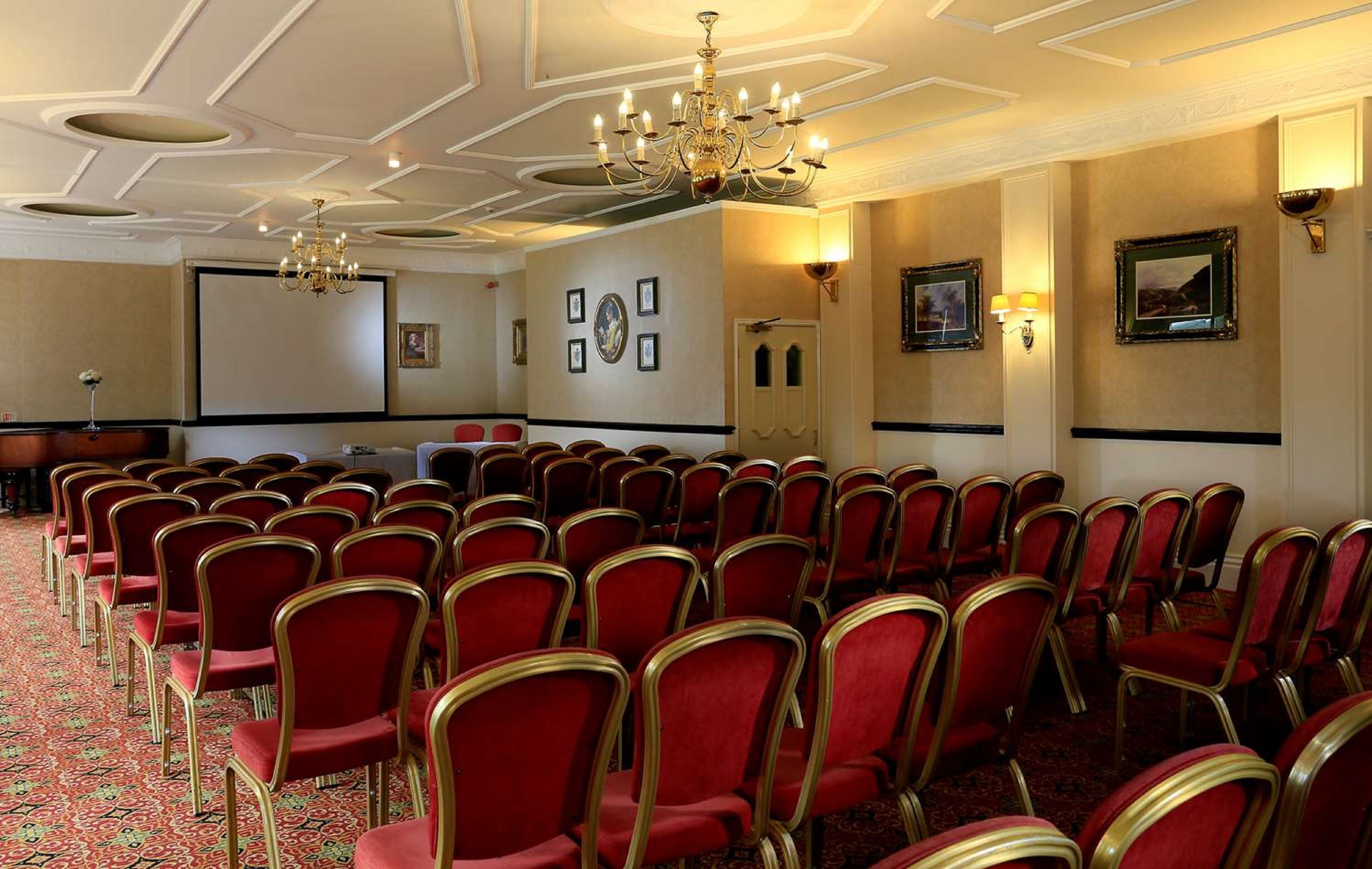 A conference room with rows of red chairs, a chandelier, framed pictures on the walls, and a carpeted floor.