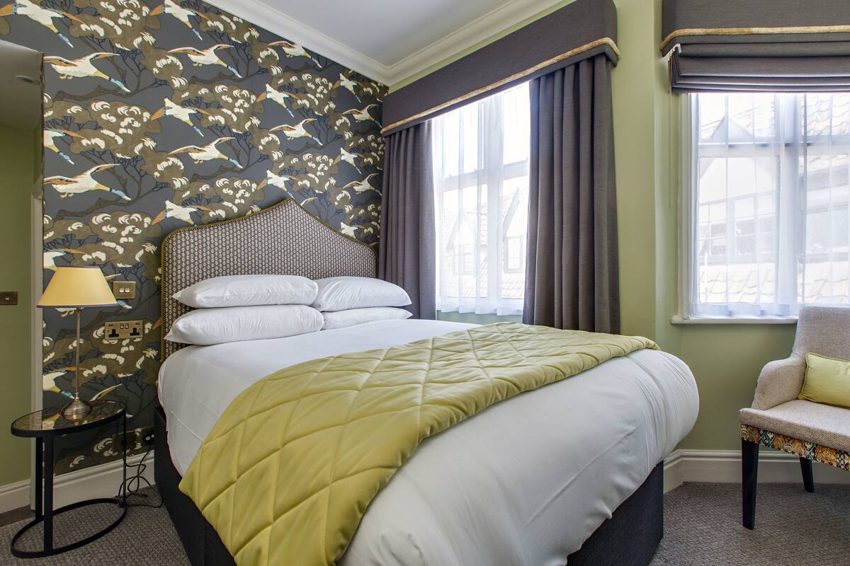 A neatly made bed with a quilted cover, a chair with a cushion, a lamp on a side table, and a window with curtains.