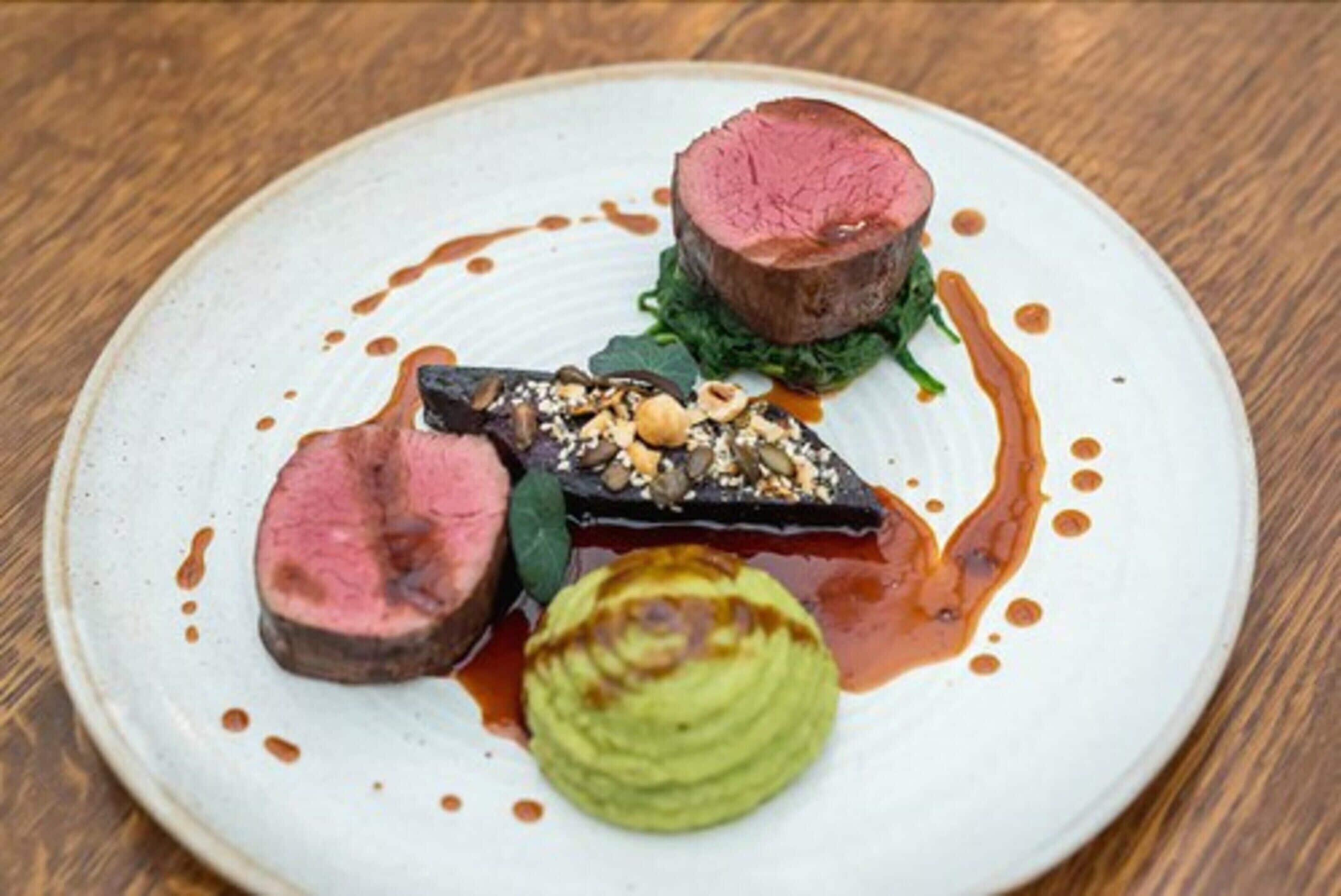 A plate with a steak, a slice of eggplant, and a green vegetable, possibly avocado, on a white plate with sauce.