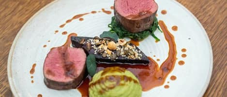 A plate with a steak, a slice of eggplant, and a green vegetable, possibly avocado, on a white plate with sauce.