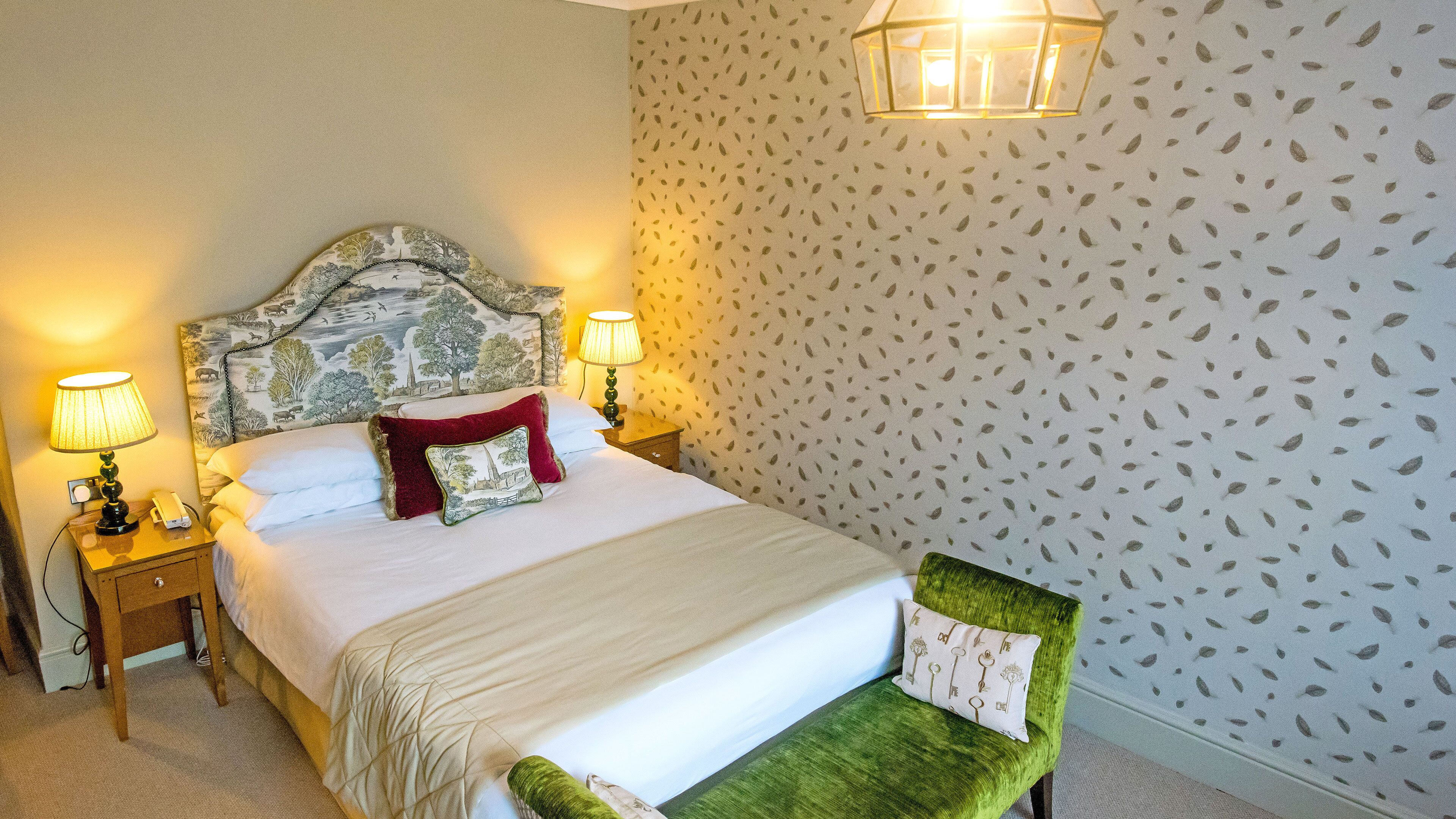 Habitación de hotel ordenada con una cama, dos mesitas de noche con lámparas, una silla verde y un papel tapiz estampado.
