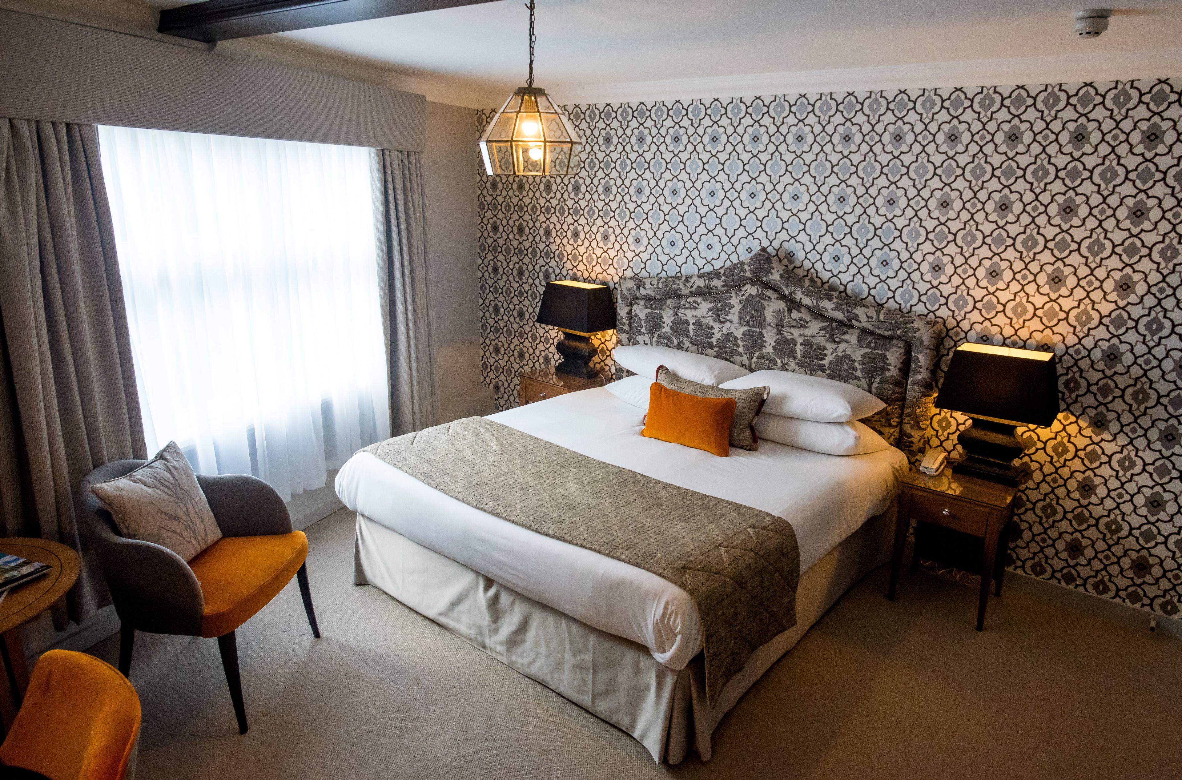 Ein Hotelzimmer mit einem großen Bett, zwei Nachttischen mit Lampen, einem Sessel und Blick ins Freie durch ein Fenster mit Vorhängen.