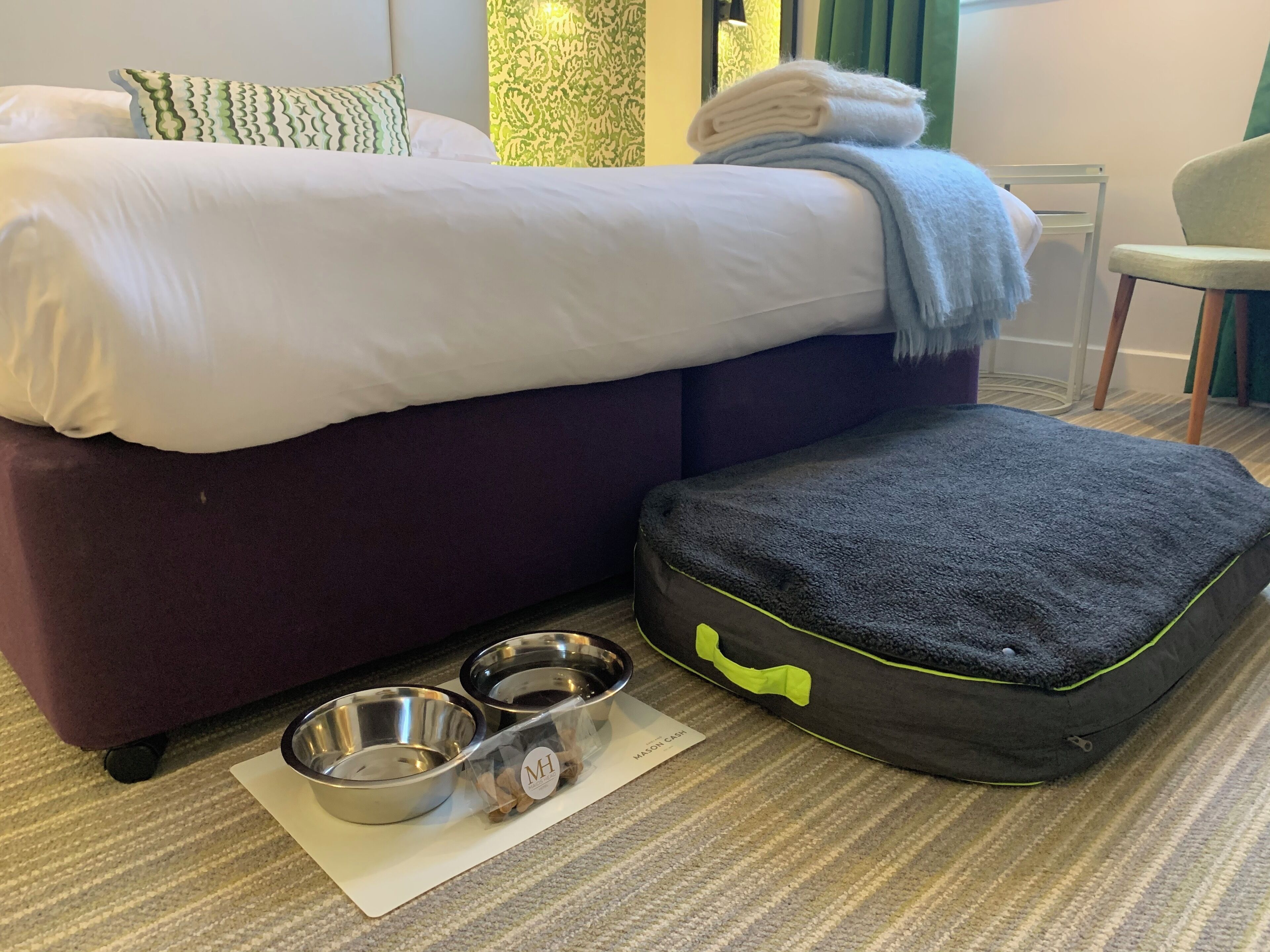 Una cama bien hecha con almohadas blancas y verdes, una toalla doblada y una cama para mascotas negra con correa. Dos cuencos metálicos están sobre un tapete junto a la cama.