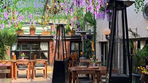 An outdoor seating area with wooden tables and chairs, surrounded by potted plants and hanging flowers.