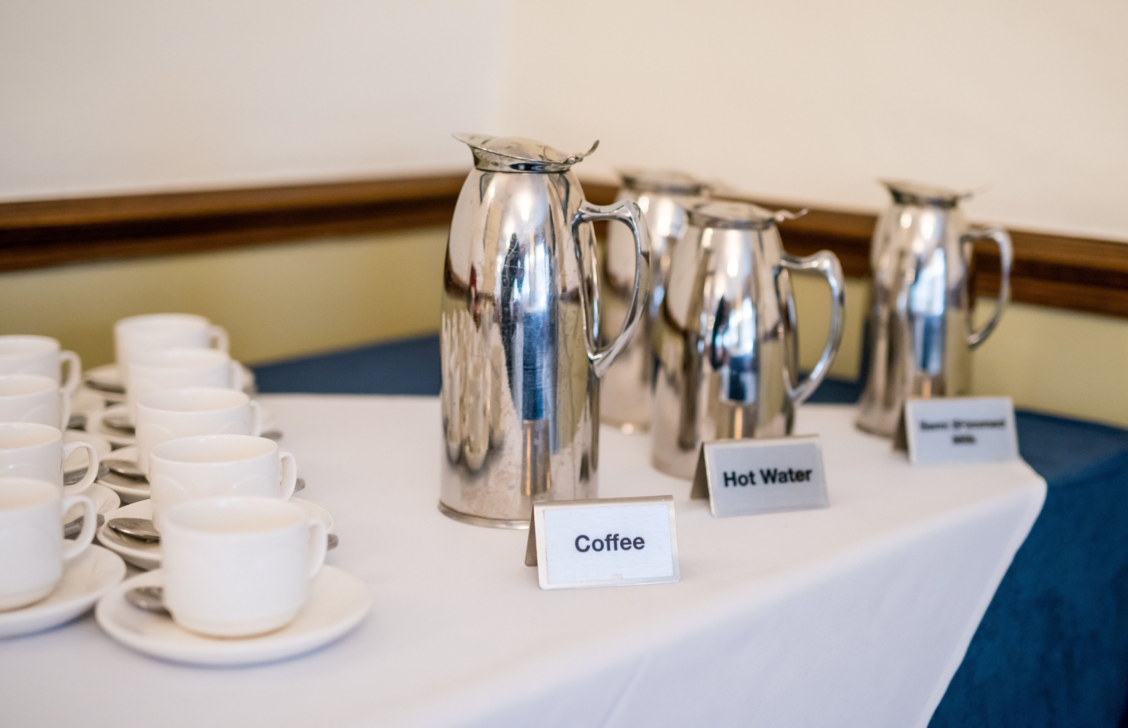 Een tafel met koffie- en heetwaterautomaten, aanduidingen en witte kopjes en schotels.
