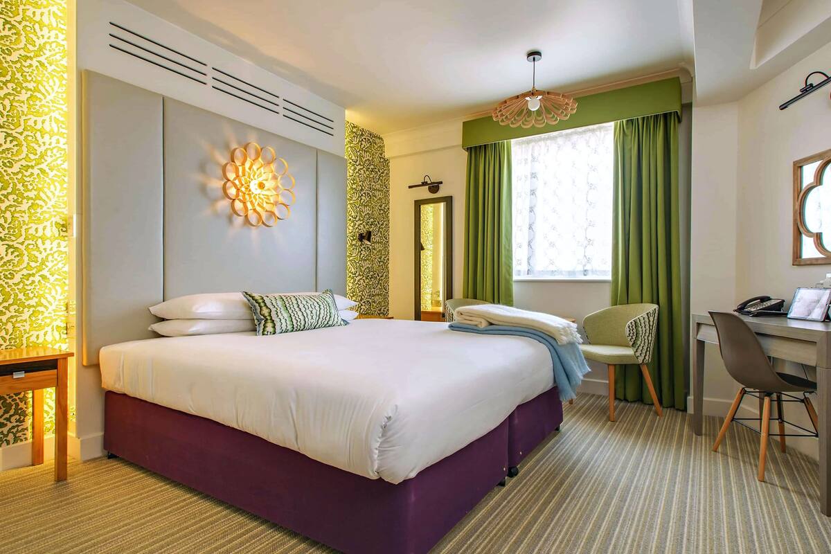 A hotel room with a large bed, a desk with a chair, a mirror, and a window with curtains.