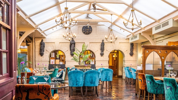 A spacious, well-lit dining area with high ceilings and chandeliers, featuring a variety of seating arrangements and a central decorative plant.