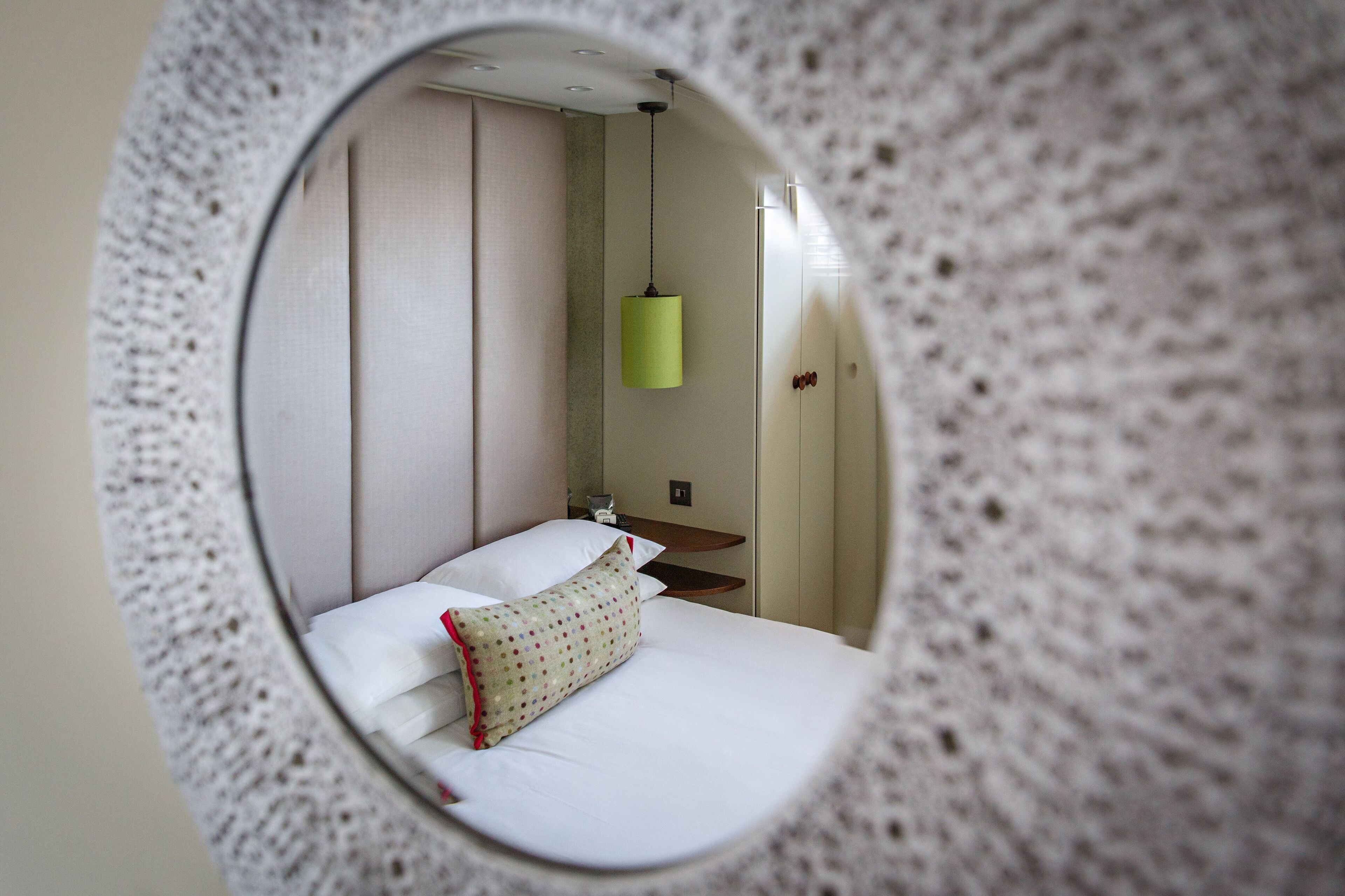 A circular mirror reflecting a bed with white bedding and a polka-dotted pillow, a green hanging light fixture, and a wooden nightstand.