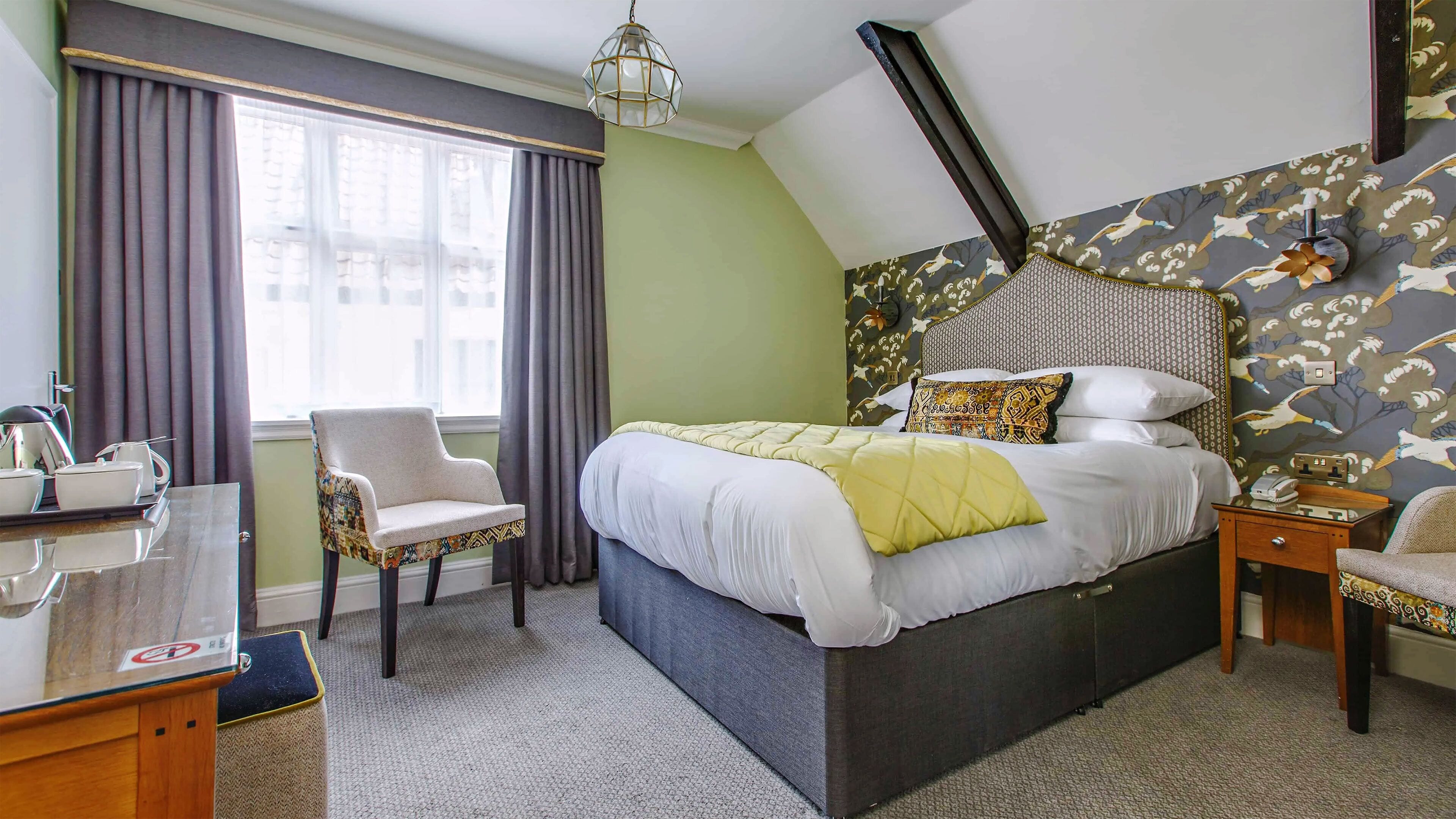 A bedroom with a large bed, a chair, a desk with a tray of items, and a patterned wall.