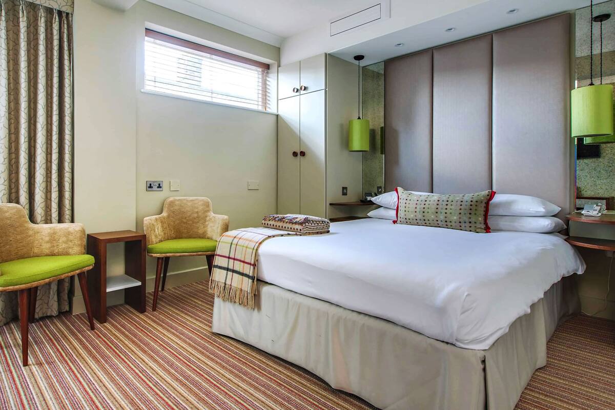 A hotel room with a large bed, two armchairs, a small table, a window with blinds, a mirror, and a green pendant light.