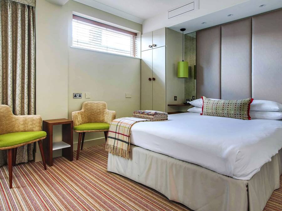 Habitación de hotel con una cama grande, dos sillones, una mesita, una ventana con persianas, un espejo y una lámpara colgante verde.