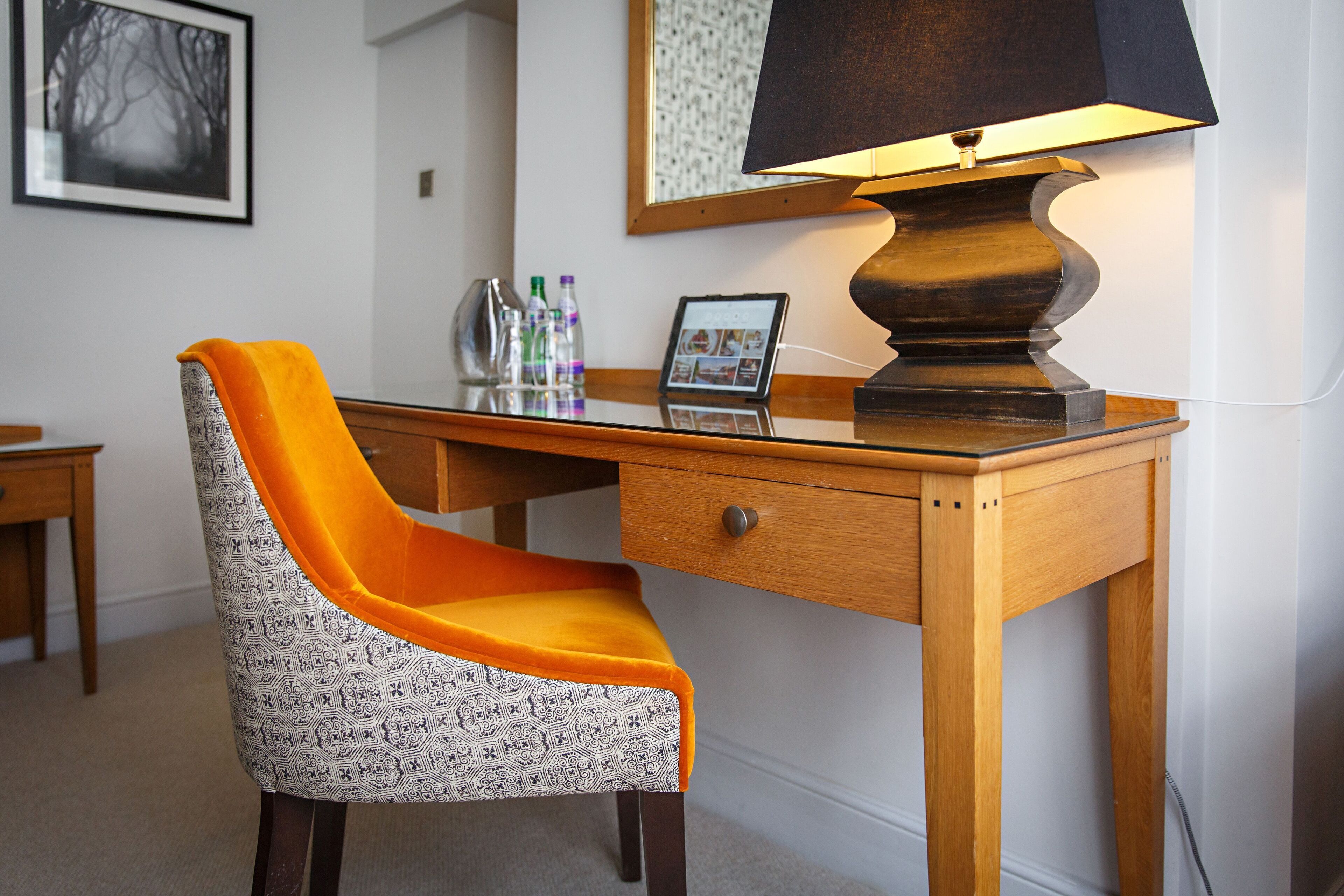 A wooden desk with a lamp, a patterned chair, a framed picture, and a tablet on the desk.