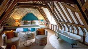 An attic bedroom with a sloped ceiling, a bed with multiple pillows, a round ottoman, a vintage bathtub, and a small table with a lamp.