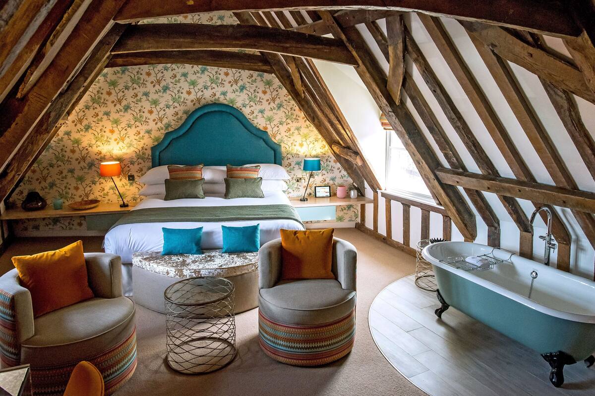 An attic bedroom with a sloped ceiling, a bed with multiple pillows, a round ottoman, a vintage bathtub, and a small table with a lamp.