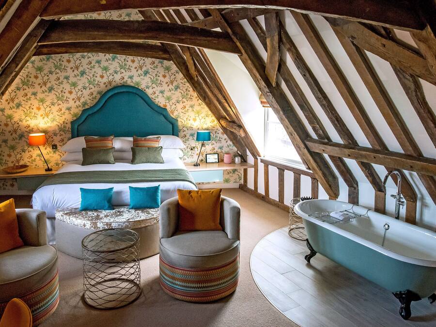 Ein Schlafzimmer im Dachgeschoss mit schrägen Wänden, einem Bett mit vielen Kissen, einem runden Hocker, einer antiken Badewanne und einem kleinen Tisch mit einer Lampe.