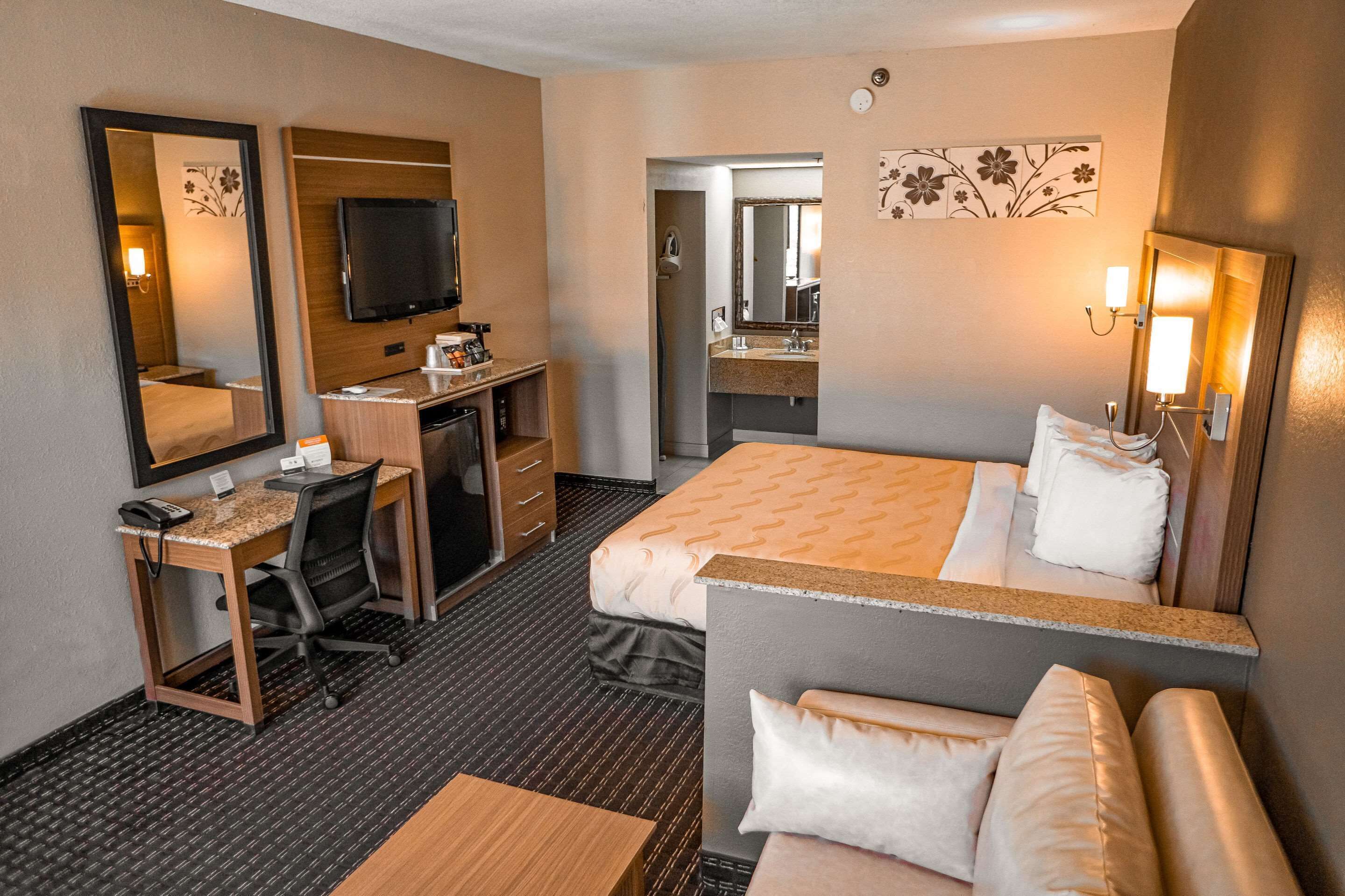 A hotel room with a bed, a desk with a chair, a television, a bathroom with a mirror, and a small table.