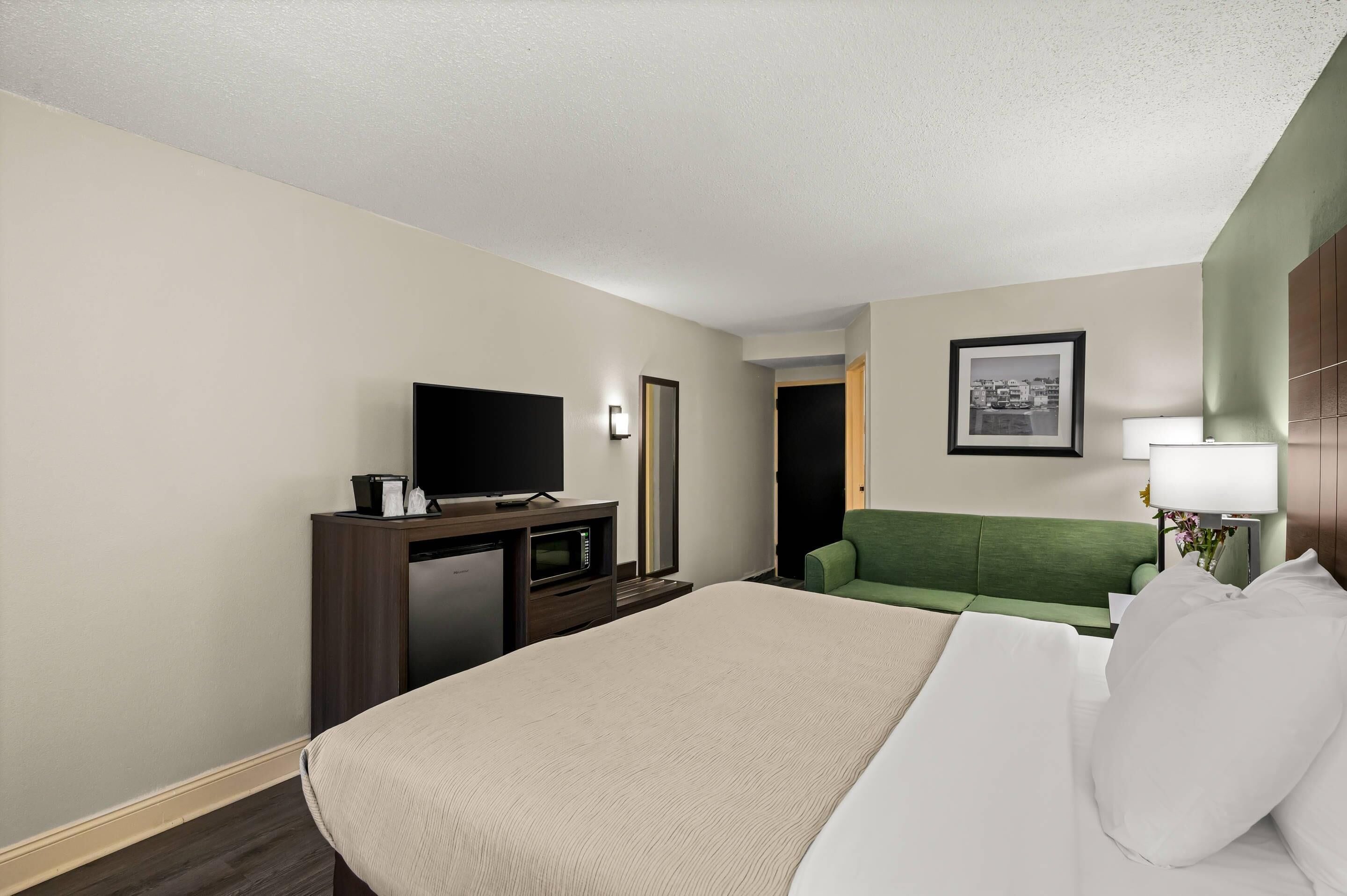 A hotel room with a bed, a desk with a television, a green sofa, a framed picture on the wall, and a lamp.