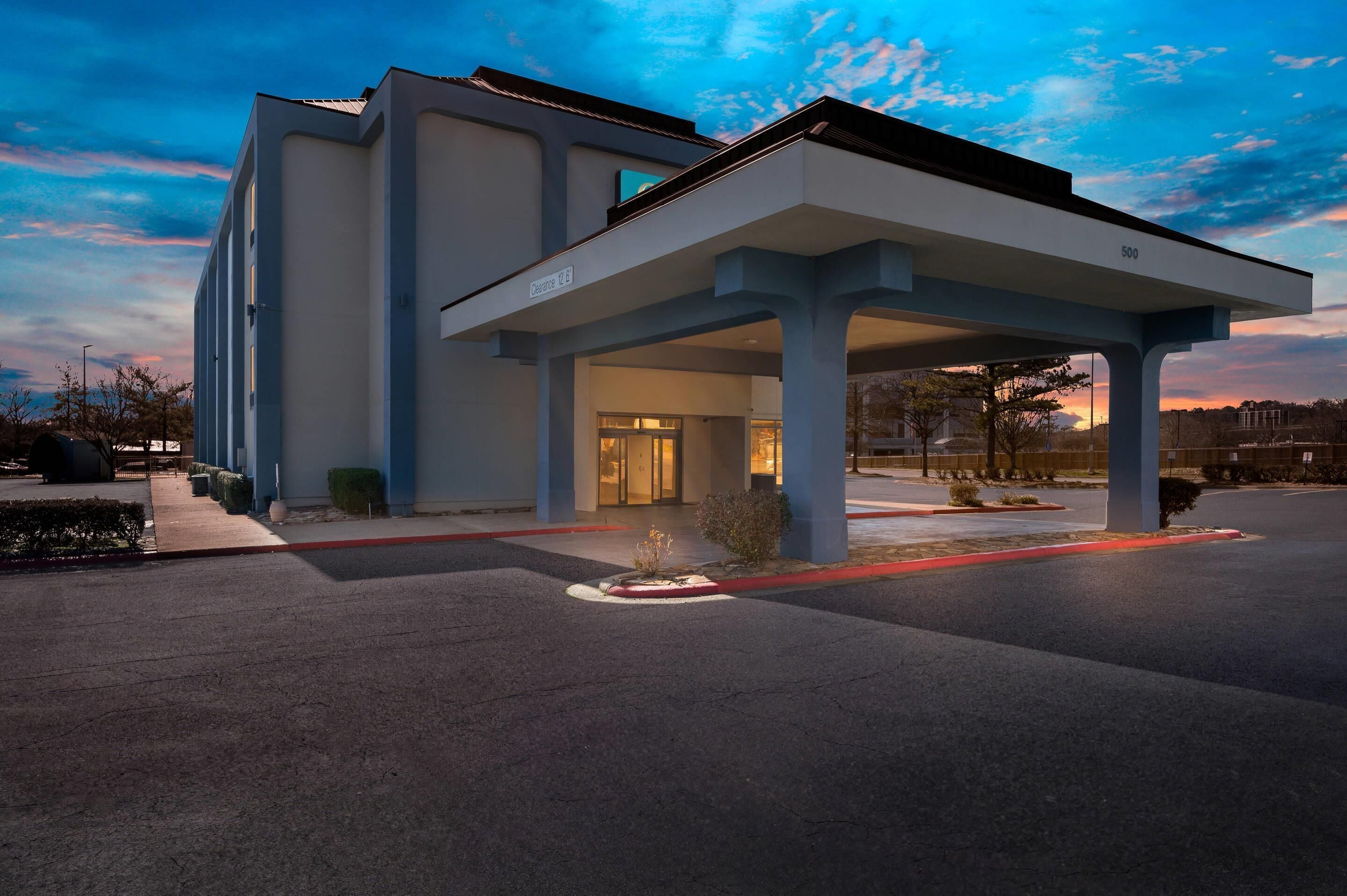 A modern hotel entrance with a covered waiting area, a clear sky, and a parking lot.