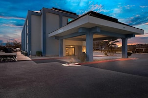 Una entrada de hotel moderna con un área de espera cubierta, cielo despejado y aparcamiento.