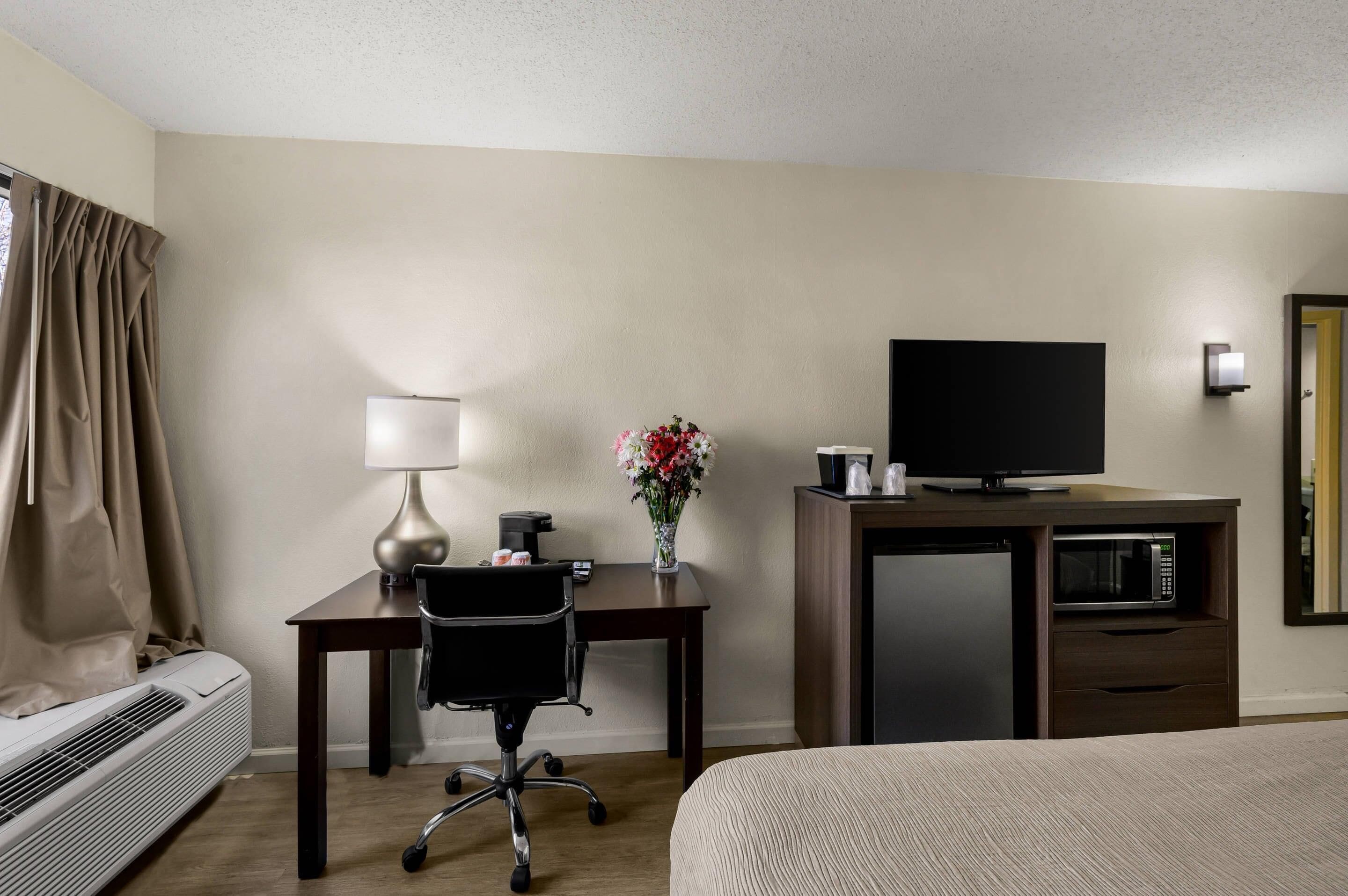 A hotel room with a bed, a desk with a chair, a television, a coffee maker, a microwave, and a vase with flowers.