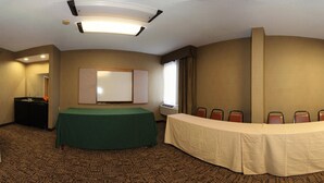 Une salle de conférence avec une table verte, une table blanche et une rangée de chaises. Un grand miroir est accroché au mur, et il y a une fenêtre avec des rideaux.