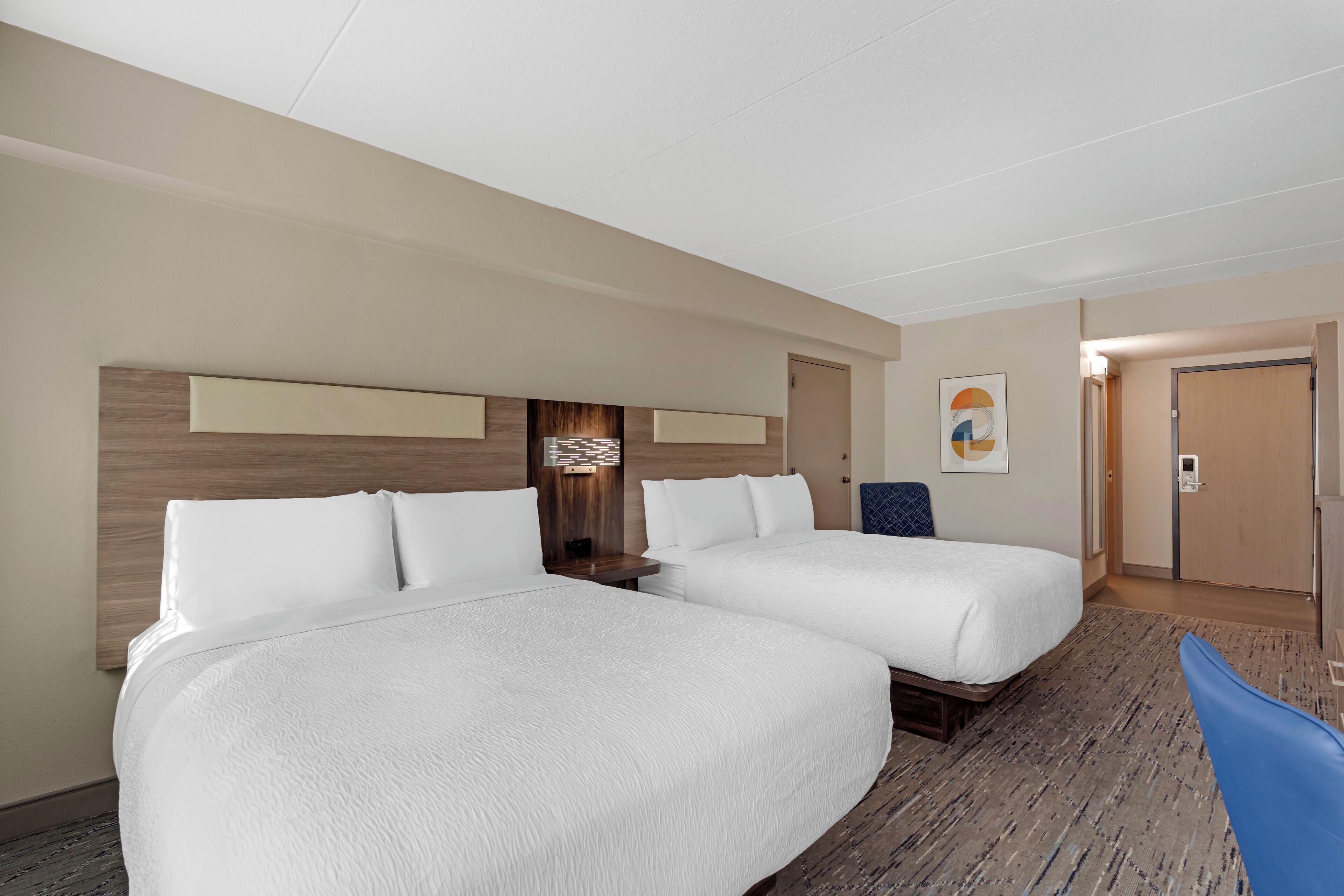 Habitación de hotel con dos camas, cabecera de madera, lámpara de pared y una puerta.