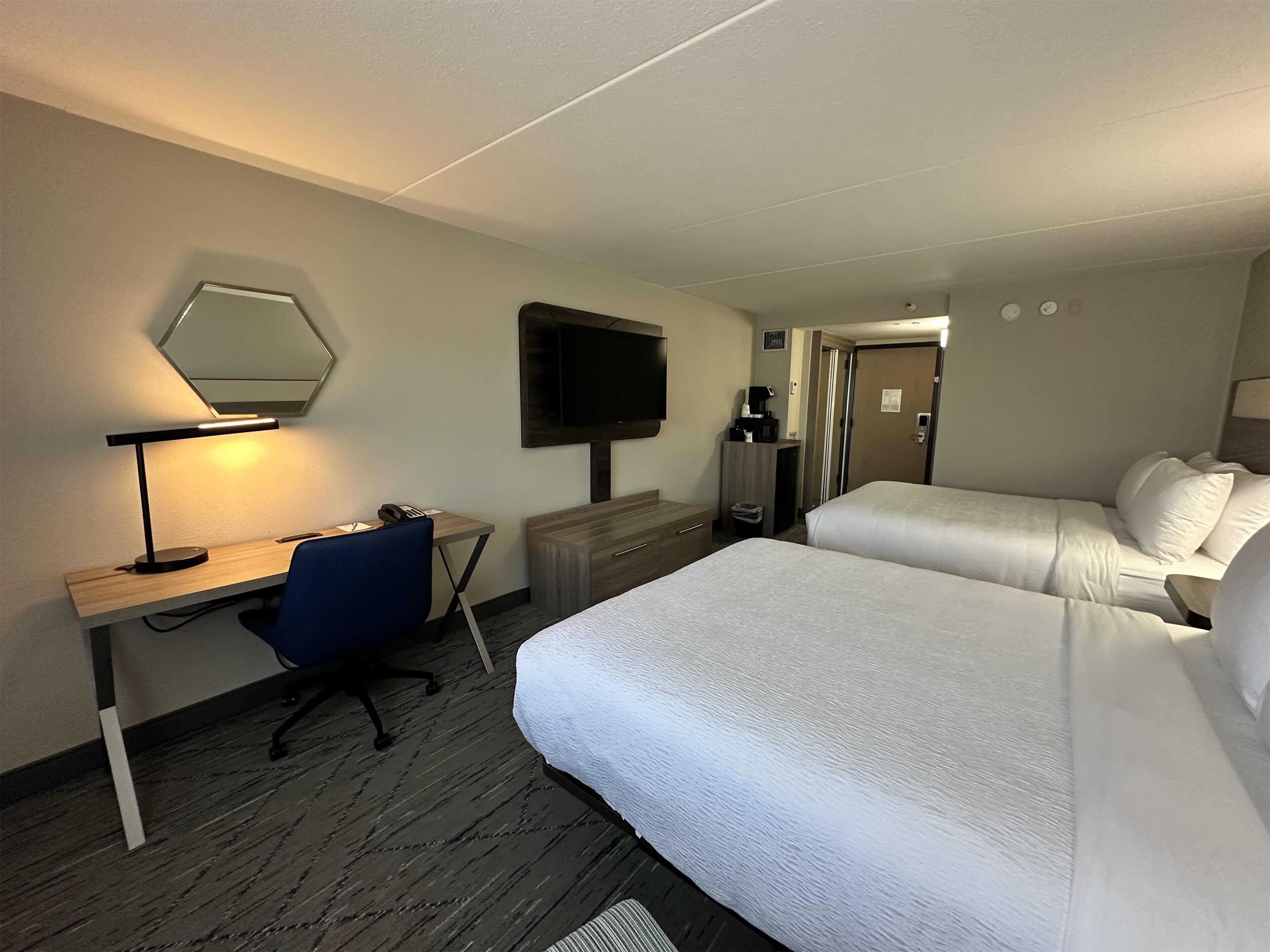 A hotel room with a desk, chair, two beds, a TV, and a mirror.