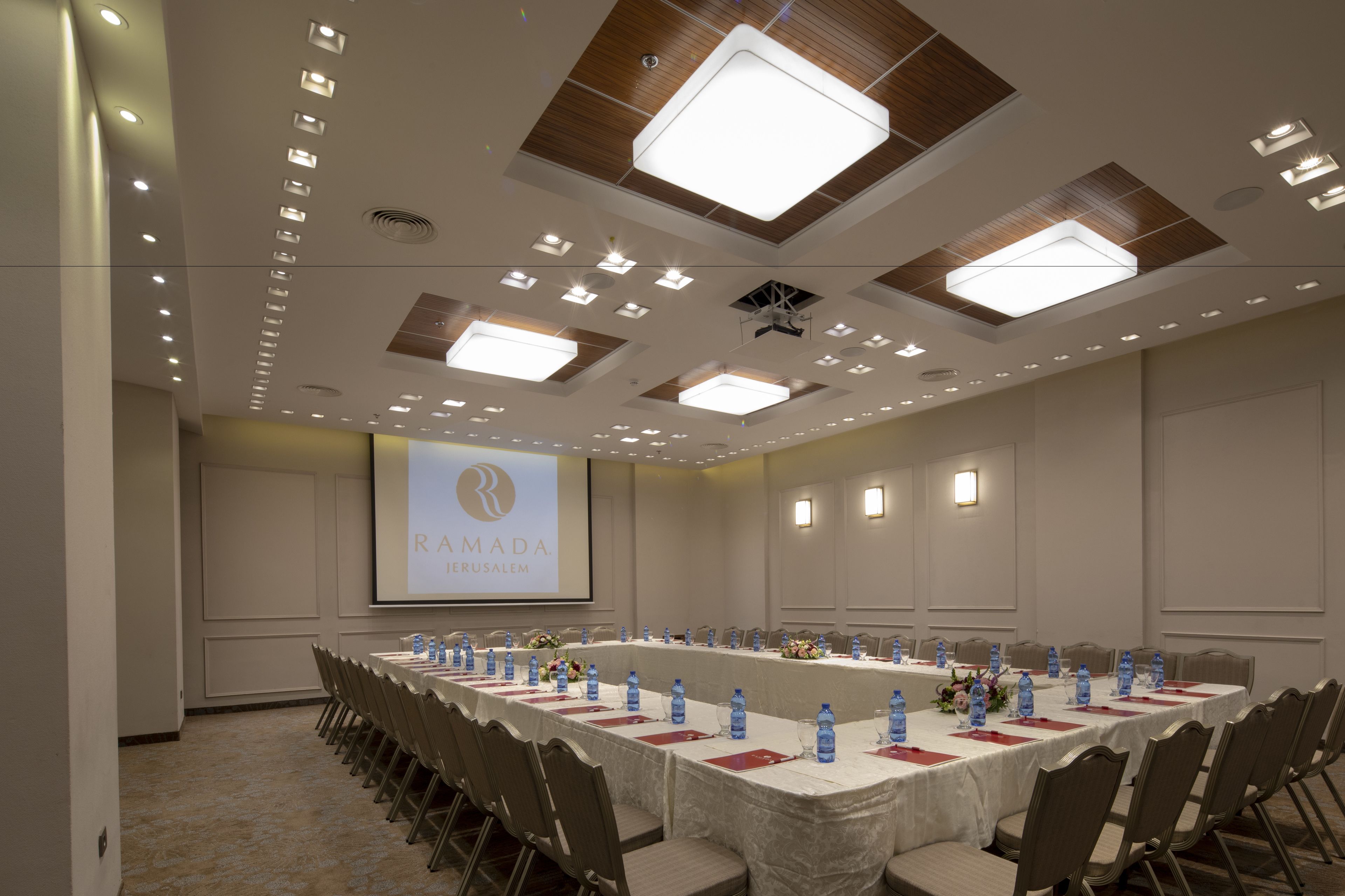 A conference room with a long table, chairs, water bottles, and a projector screen displaying "RAMADA Jerusalem".