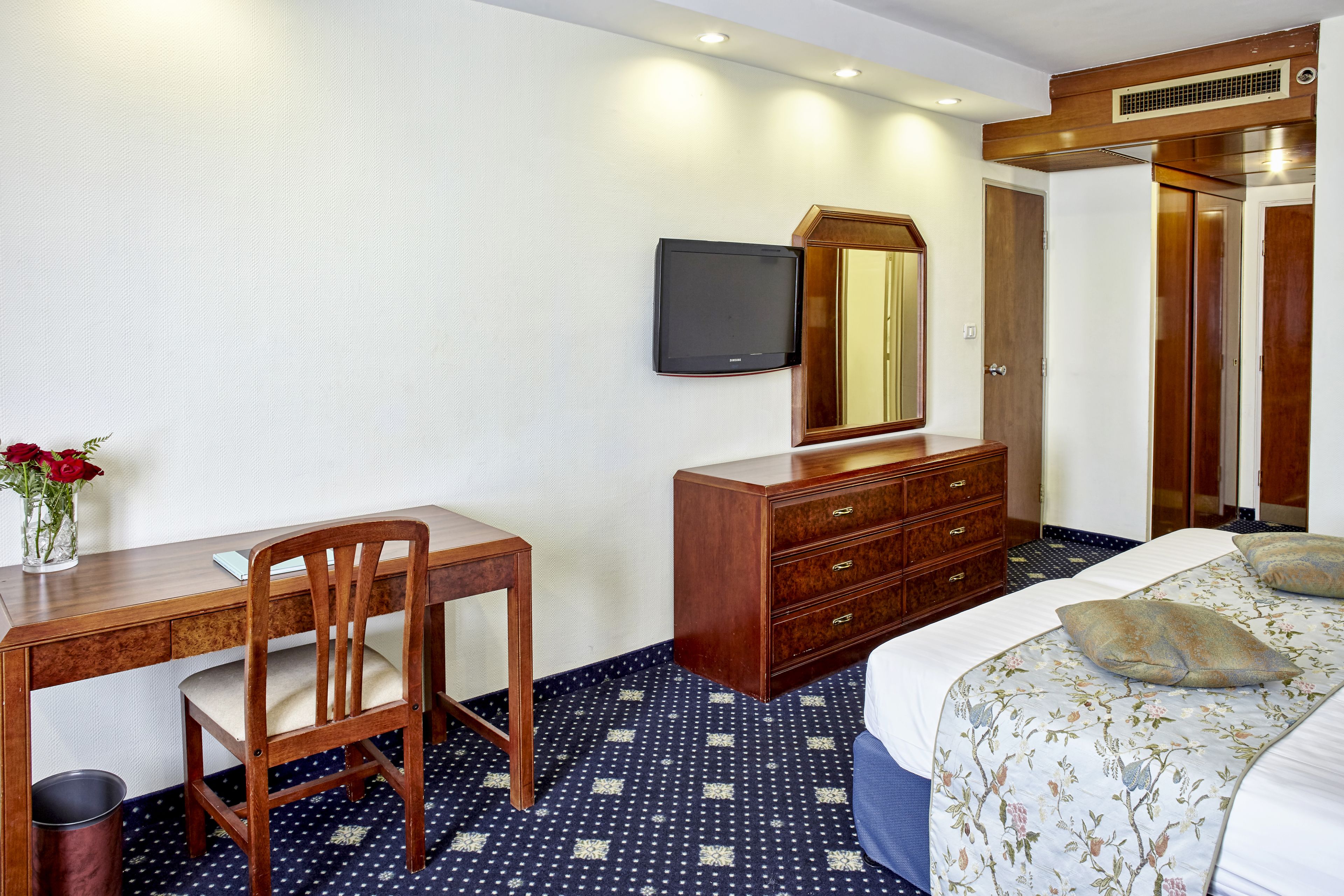 A hotel room with a wooden desk, a chair, a dresser, a bed with floral bedding, a television, and a mirror.