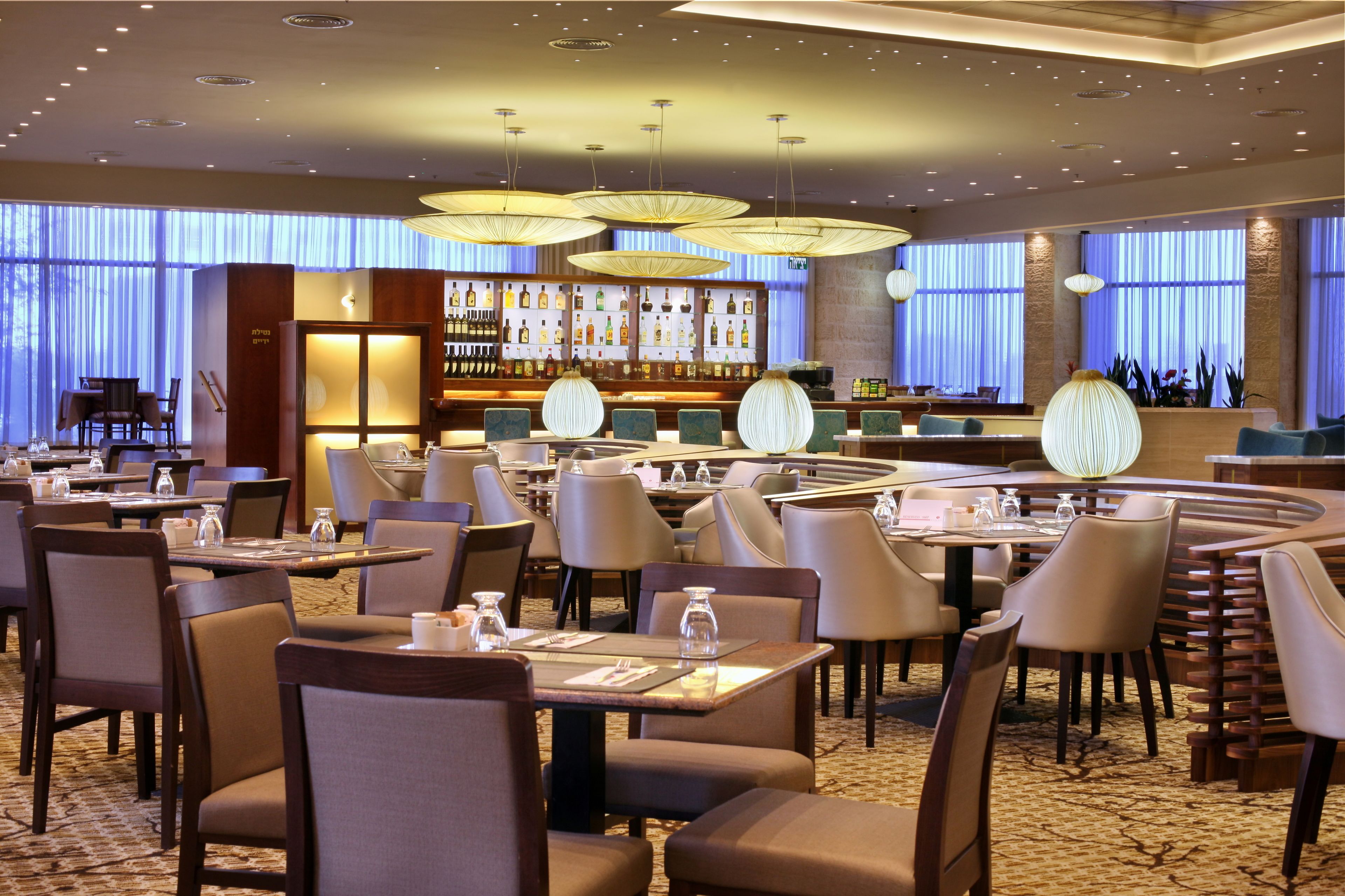 A hotel lobby with multiple dining tables and chairs, a bar area with a variety of bottles, and large windows with curtains.