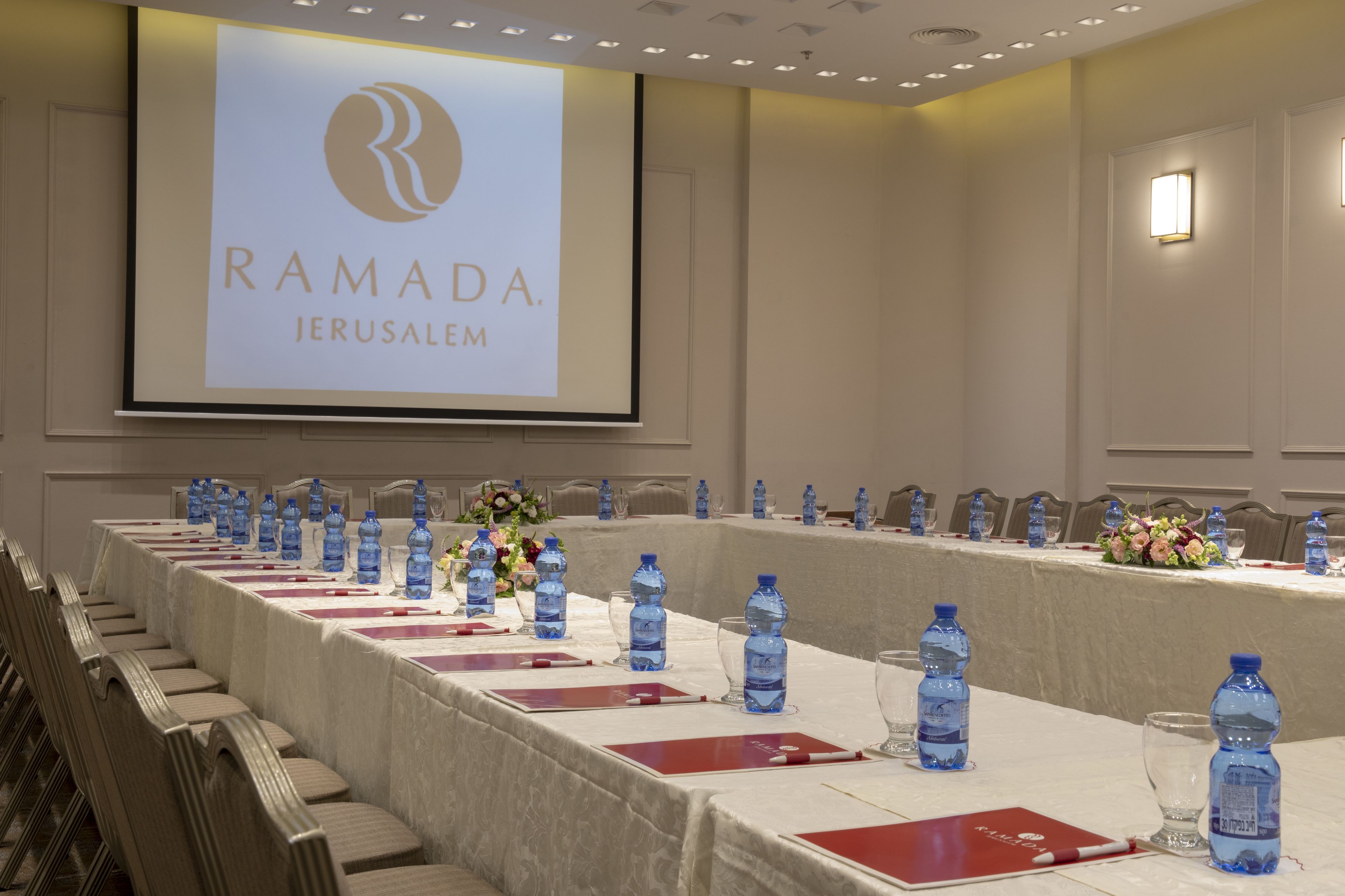Een vergaderruimte met een lange tafel, waterflesjes en rode mappen. Een scherm toont 'RAMADA JERUSALEM'.
