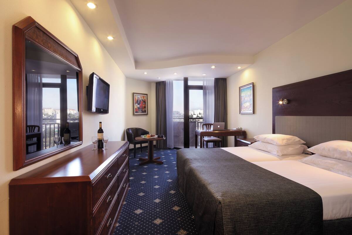 Quarto de hotel com uma cama grande, uma escrivaninha com um laptop, uma cadeira, uma televisão e vista para a cidade pela janela.