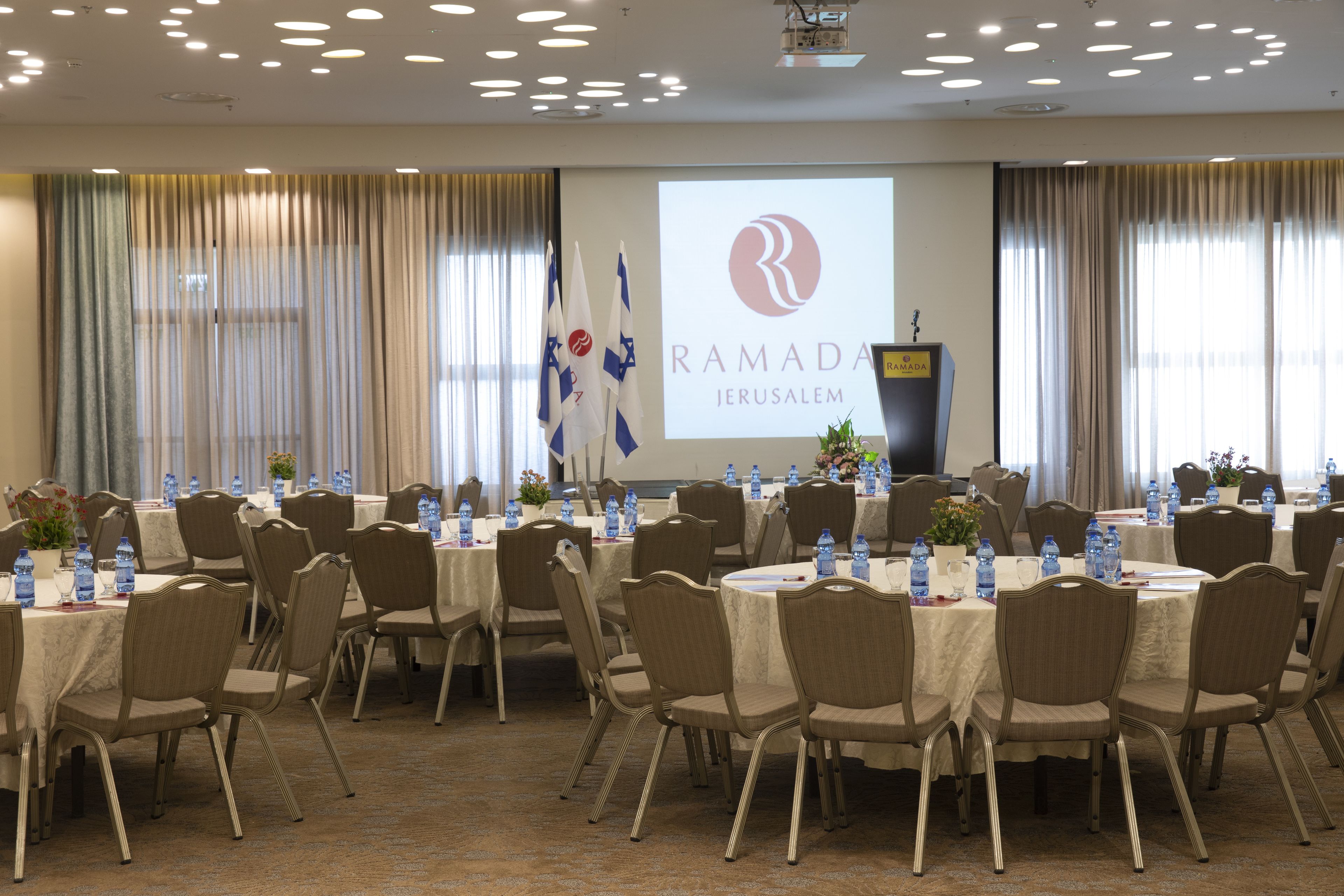 A conference room with round tables, chairs, and a projection screen displaying the Ramada Jerusalem logo.